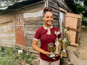 Andreina, a teenage girl, displays her music trophies while standing in from of her small wooden home.
