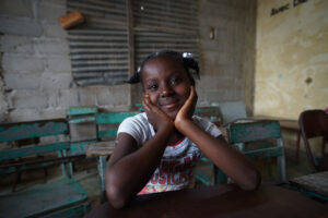 A Haitian girl sits at a desk in a concrete block classroom surrounded by metal chairs with peeling teal paint with chin resting on her hands and a slight smile 