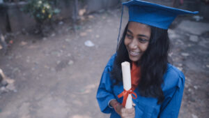 A smiling girl with long black hair wears a graduation cap and gown and holds a rolled diploma tied with a red ribbon