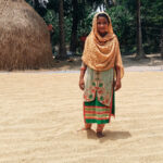 Girl in Bangladesh wearing colorful headscarf and kurta stands amidst grain in front of hay.