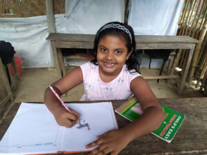 A young girl dressed a pink shirt sits at a rough wood table drawing in a work book and smiles at the camera. 