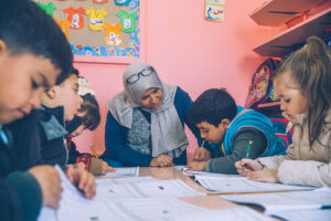 A teacher wearing a headscarf sits with a group of young children at a table in a classroom helping them read and write. 