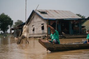 A boy in a rough wood fishing boat paddles past a floating shanty in Cambodia