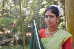 A girl in traditional dress holds a Bangladesh flag in front of a forrested background