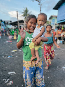 A young girl holding a baby in an impoverished neighborhood in teh Philippines waves to the camera