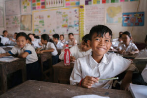 A smiling boy in a classroom filled with children sits at a wooden desk and looks up in thought. 
