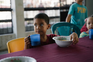 Boy holding a cup with a bowl of food at a lunch table. 