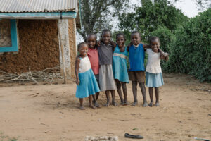 Six small barefoot children stand arm in arm in front of a mud house