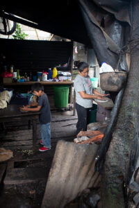 A boy and his mother prepare tortillas in an open air kitchen made of rough board walls and floor and corrigated metal attached to a tree
