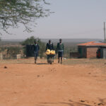 3 teen boys walk through an arid village toward the camera, one pushing a wheelbarrow full of jerrycans of water