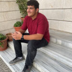 A teen boy smiling with confidence wearing a red school uniform shirt and black trousers sits on some marble steps out side a building