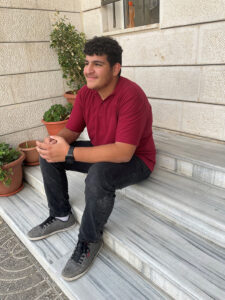 A teen boy smiling with confidence wearing a red school uniform shirt and black trousers sits on some marble steps out side a building