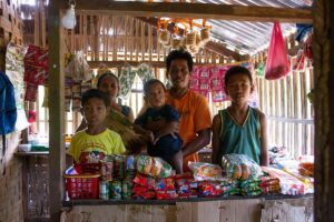 A family of mother, father holding baby, and two sons stand behind a makeshift counter in a small structure of wood and woven matting. On the counter are a variety of food packages. 