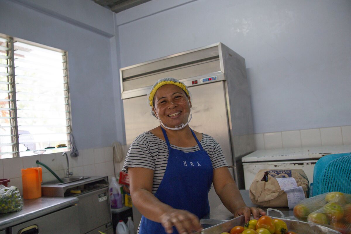 A woman in a blue apron smiles broadly as she prepares vegatables.