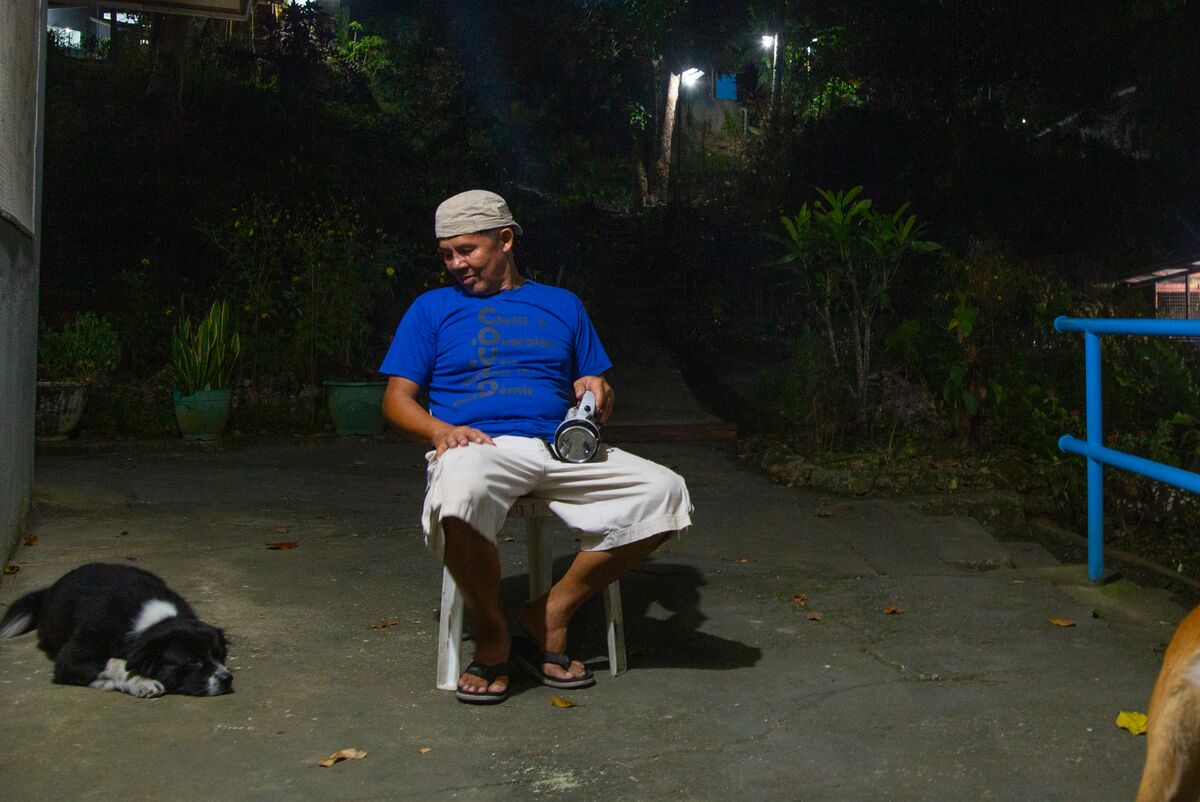 A man in a blue t-shirt and white cargo shorts sits on a plastic chair holding a flashlight. A medium shaggy black dog lays nearby. 