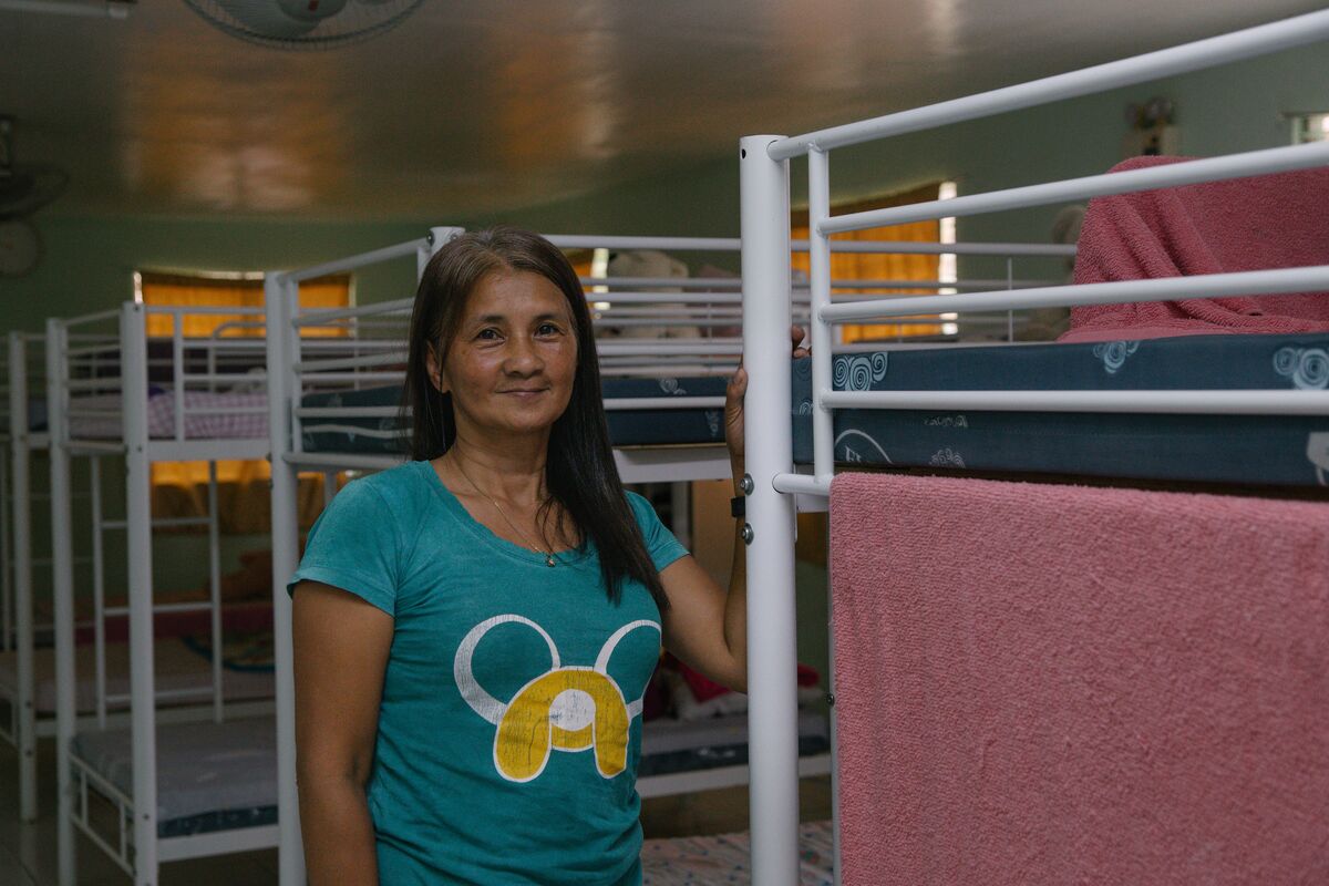 A middle-aged Filipino woman with long hair stands next to bunkbeds in a dormitory. 