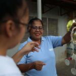 A woman in with glasses in a blue polo shirt holds up a windchime made of shells.