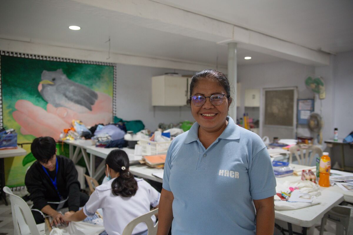 Ate Teresa in a light blue polo and glasses stands in the craft room with teens doing crafts in the background.