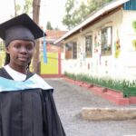 A woman in a black cap and gown stands outside a schoolhouse. She is smiling at the camera