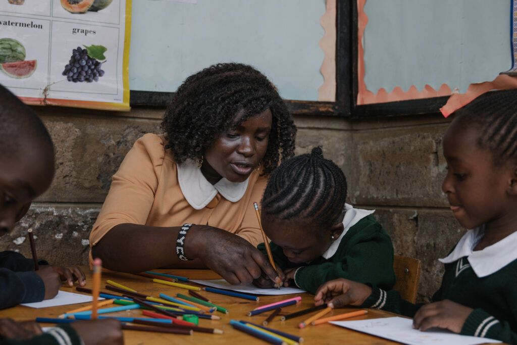 A woman helps a young student write with a pencil. There are colored pencils on the desk in front of them.