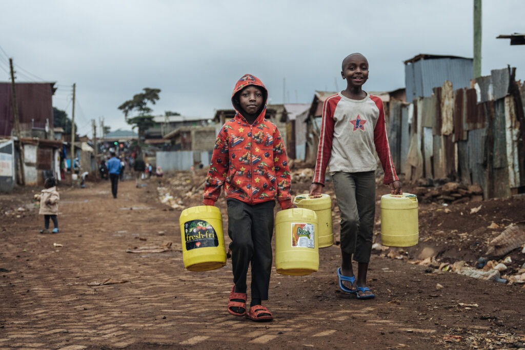 Two kids carry yellow jugs of water through an impoverished community in Nairobi, Kenya. They are smiling.