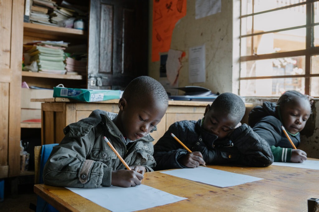 Three kids sit at a classroom table. They are writing on paper with pencils.