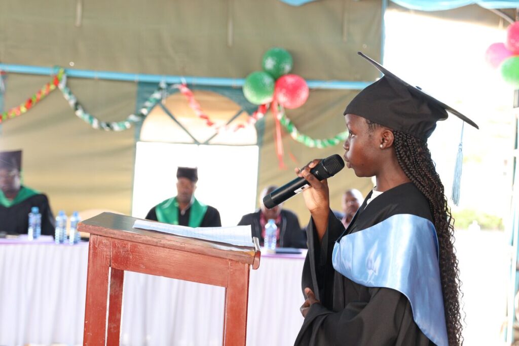 A young woman in a graduation cap and gown speaks into a microphone at a podium
