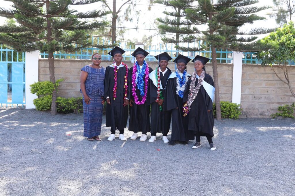 Five young adults wear black graduation gowns and caps and colorful garlands around their necks. They are standing outdoors next to a woman wearing a blue dress.