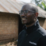 Jacob wearing glasses and a black onechild jacket stands in front of a brick house with a metal roof.